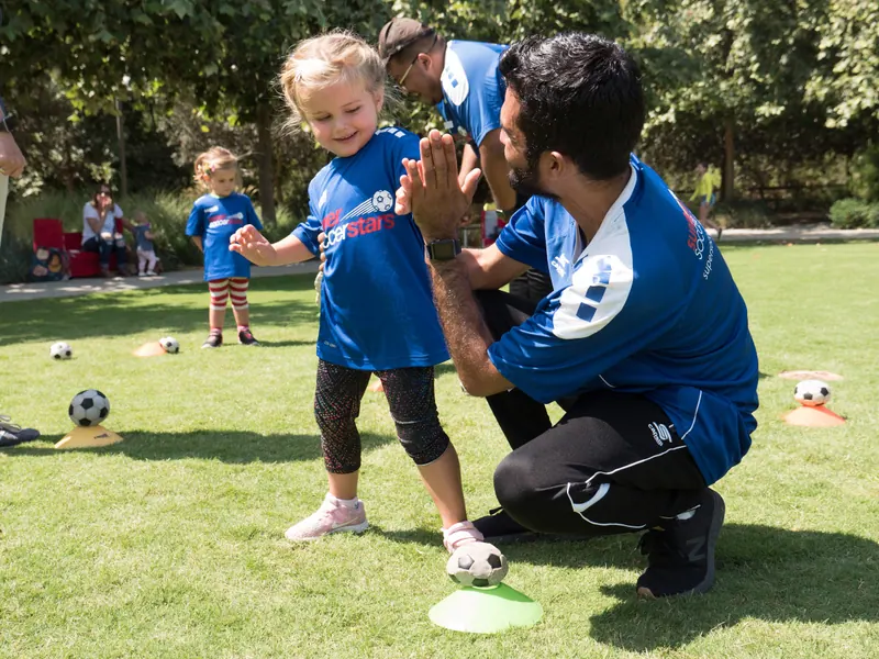 A coach crouches to share a high-five with a young player on a sunny field
