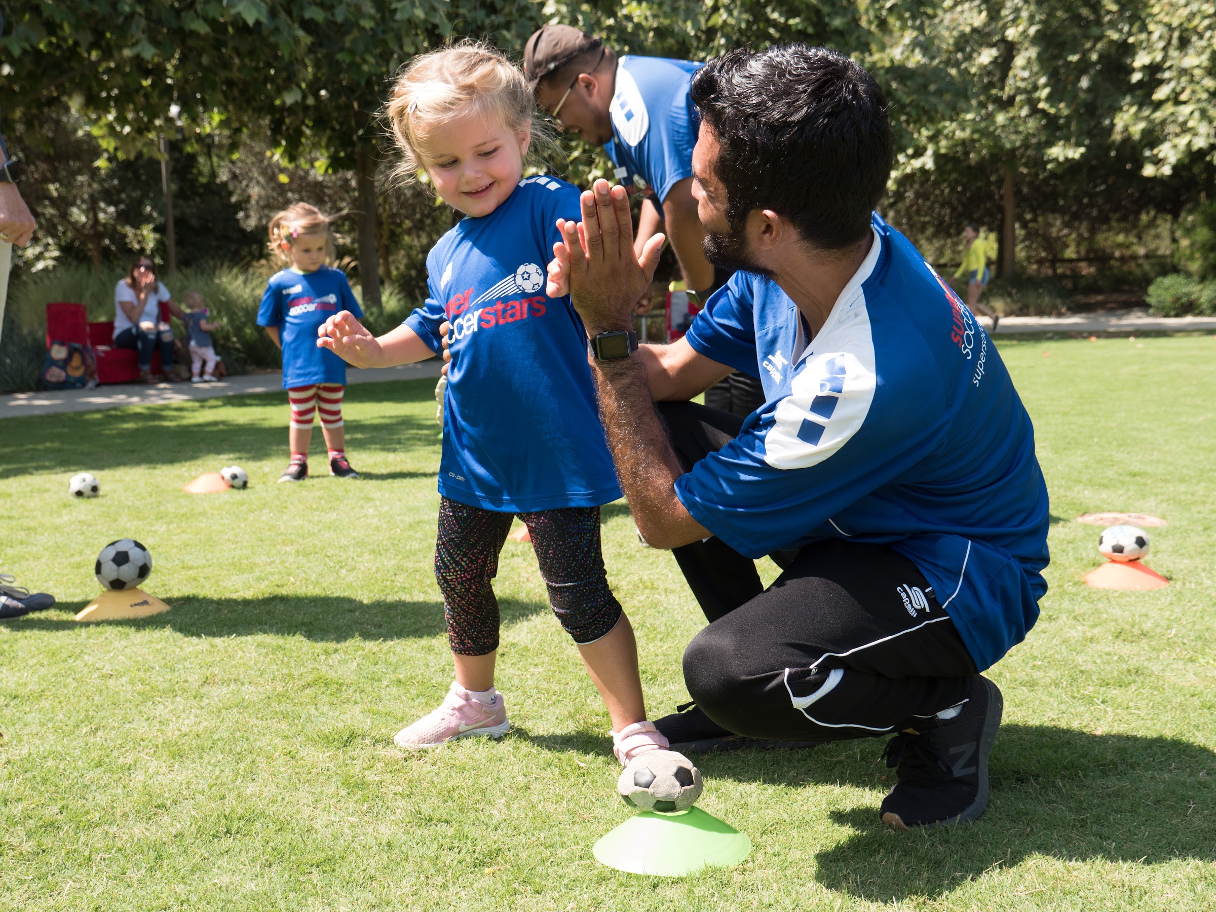 A coach crouches to share a high-five with a young player on a sunny field