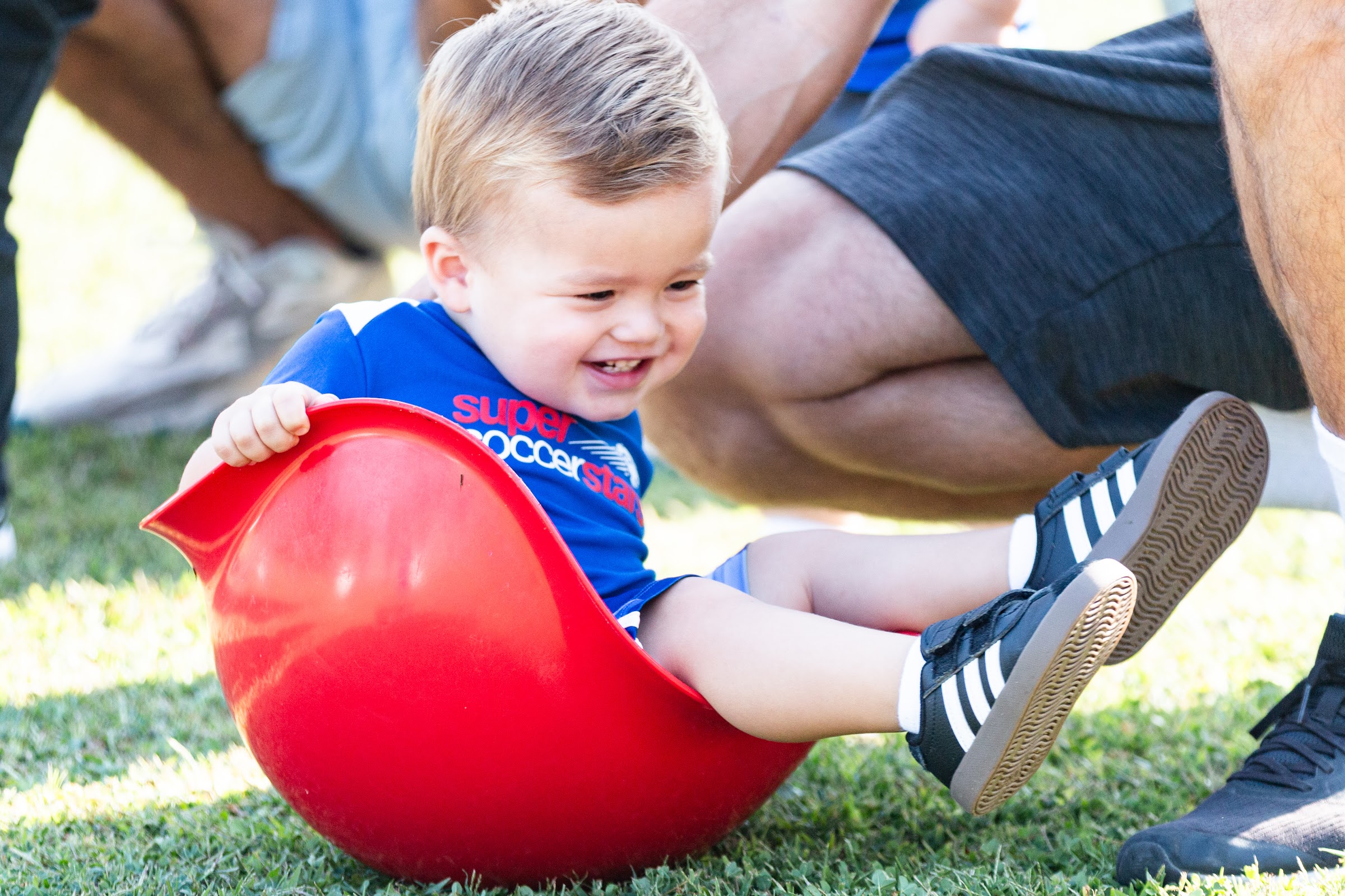 A toddler laughs while playing outdoors with a caregiver nearby